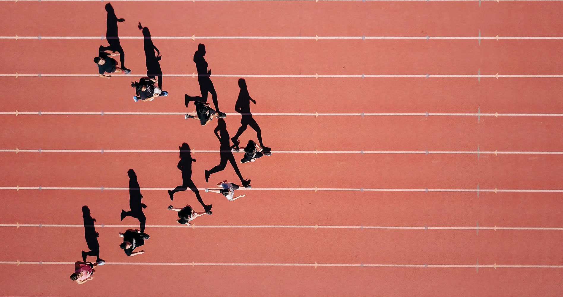 Een foto van een atletiekbaan, van bovenaf genomen. Acht personen lopen op de baan en vormen een driehoek met de punt naar rechts. Hun schaduwen vallen mooi op de baan, wat de illusie wekt dat ze de Progress-vlag uitbeelden.